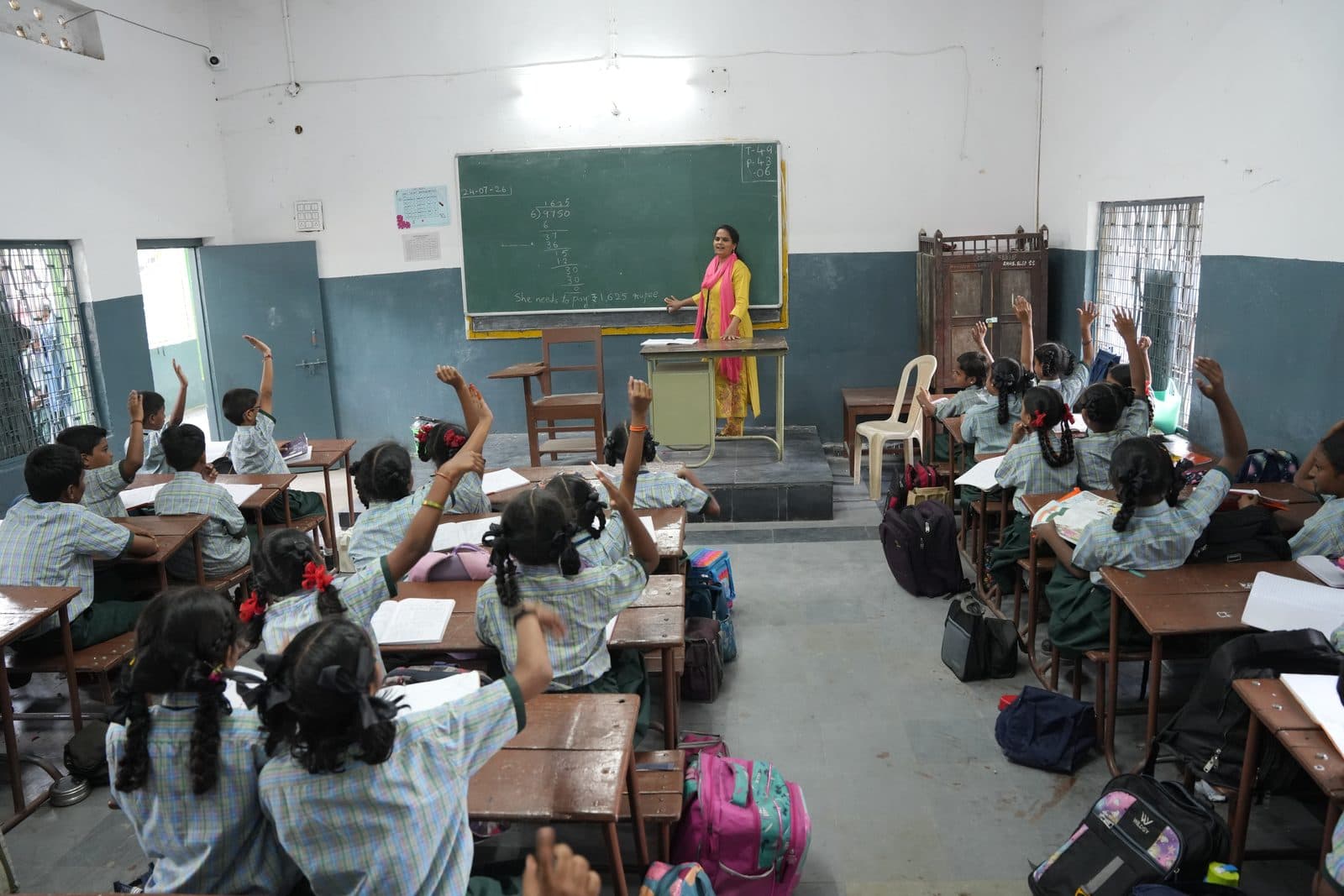 Primary students raising their hands to answer as the teacher works at the blackboard