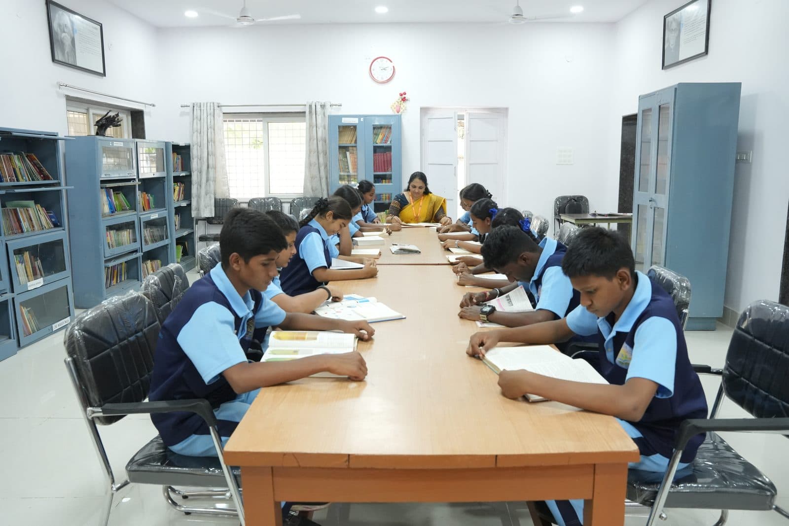 Students seated along a long library table reading books, with the librarian at the far end